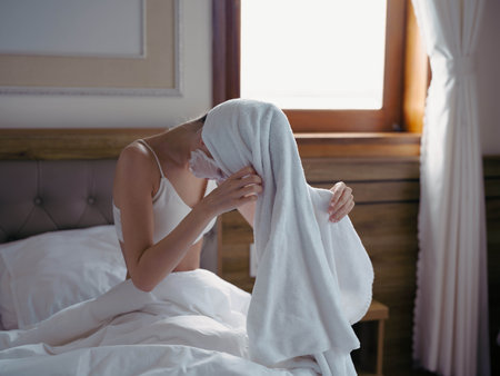Young woman lying on bed with moisturizing beauty face mask and white towel on her head after shower, lifestyle home skincare, smile.の写真素材