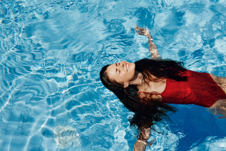 Happy woman swimming in the pool in red swimsuit with loose long hair relaxed in the sunshine, skin protection with sunscreen, concept of relaxing on vacation in tropical climate, lifestyle.の写真素材