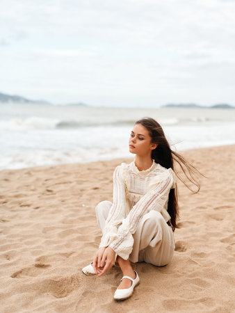 Serene Beach Beauty: A Young Woman Enjoying the Sea Breeze in a Casual White Dress, Gazing at the Blue Ocean on a Sunny Summer Dayの写真素材