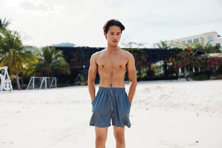 Happy Asian Man Enjoying Vacation on a Tropical Beach, Smiling and Showing Muscular Torso in Shirtless Portraitの写真素材