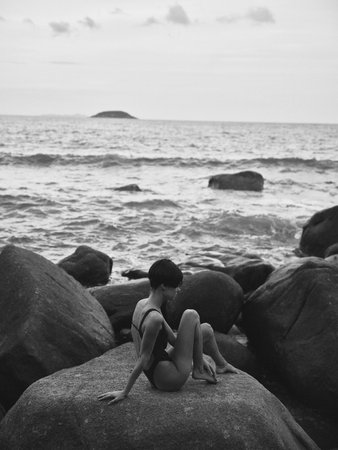 Solitude in Sunset: Lonely Silhouette of a Sad Woman Relaxing on a Stone by the Ocean, Looking out at the Horizonの写真素材