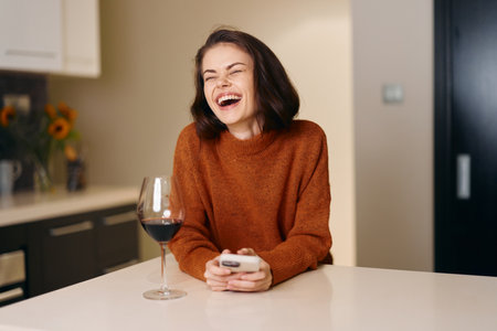 Happy Young Woman Enjoying a Glass of Wine, Sitting on a White Sofa and Holding a Mobile Phoneの写真素材