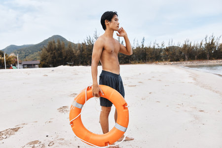 Lifeguard on the Beach, Summer Asian Man Saving Lives - A depiction of a diligent lifeguard in vibrant swimwear, holding a red lifebuoy, exuding a sense of duty and alertness. The background portrays a picturesque seaside with golden sand, sparkling blue oの写真素材