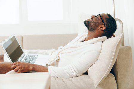 Smiling African American Man in Bathrobe Working on Laptop in Modern Home Office Or Young African American Freelancer Typing on Laptop while Relaxing on Sofa in Morning Lightの写真素材