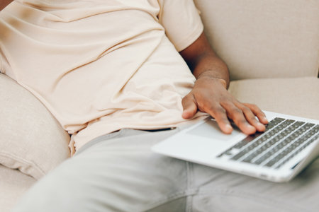 Smiling African American Man Working on Laptop in Cozy Living Room, Making a Phone Call for Online...