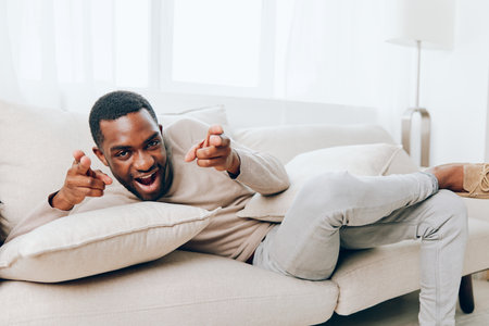 Relaxed African American Man Sitting on a Comfortable Sofa in his Living Room, Smiling and Holding a Pillow He is Enjoying a Peaceful Weekend at Home, Watching Something Online and Thinking with aの写真素材