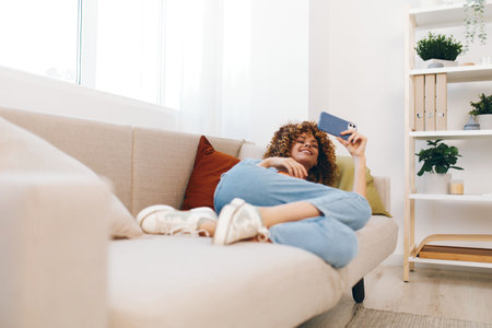 Happy woman enjoying online games and chatting on her mobile phone while sitting on a cozy sofa at home.の写真素材