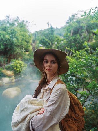 A woman wearing a hat and carrying a backpack stands near a river in the woods