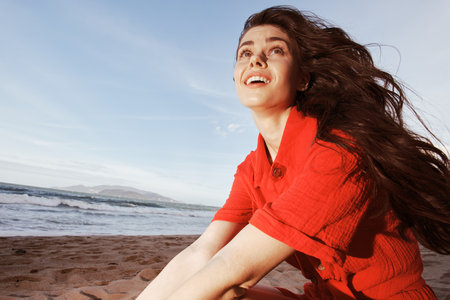 Serene Sunset: Beautiful Woman Sitting on the Beach, Enjoying Freedom and Vacation Bliss in the Warm Summer Eveningの写真素材
