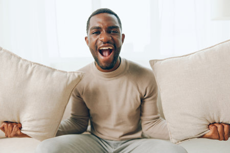 Relaxed African American man sitting on a comfortable sofa in his modern living room, smiling and holding a pillow He is enjoying a peaceful weekend at home, thinking and watching something onlineの写真素材