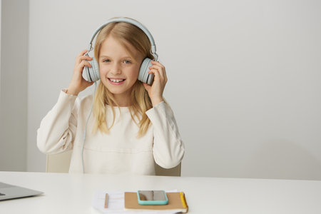 Smiling schoolgirl using a laptop for elearning and online education at home She is sitting at a desk, typing on the keyboard with headphones on With her beautiful caucasian features, the cute girlの写真素材