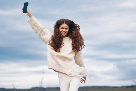Mountain Woman: Embracing Natures Freedom. Smiling Adventurous Traveler Captures a Hiking Selfie amidst Beautiful Landscape, Peak Explorer in Sweater, Spring Outdoors. Mobile Communication in Cyberspace with the Majestic Mountain Range as the Background.の写真素材