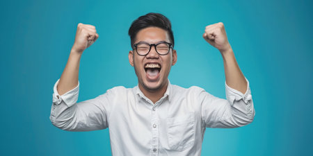 Confident Asian man in white shirt and glasses raising fist against blue background portraitの素材