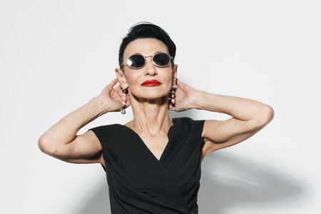 Fashionable woman in sunglasses and black dress posing with hands on head against white wall,...