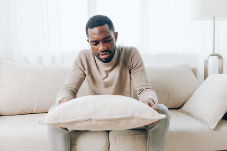 Smiling African American Man Sitting on a Modern Sofa at Home, Relaxing on the Weekend He is Holding a Pillow and Looking Relaxed and Comfortable The Living Room Background Adds to the Cozy Atmosphereの写真素材