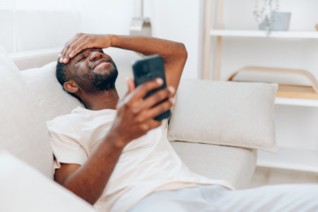 Happy African American Man Holding Smartphone and Relaxing on a Black Sofa at Home Modern Lifestyle and Technology, Enjoying Communication and Connectionの写真素材