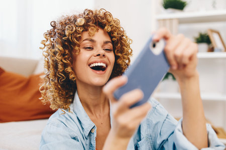 Happy woman holding mobile phone and smiling while relaxing on the cozy sofa at homeの写真素材