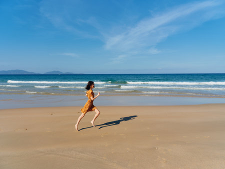 Woman in a vibrant yellow dress joyfully running along the sandy beach shore with the vast ocean in the backgroundの写真素材