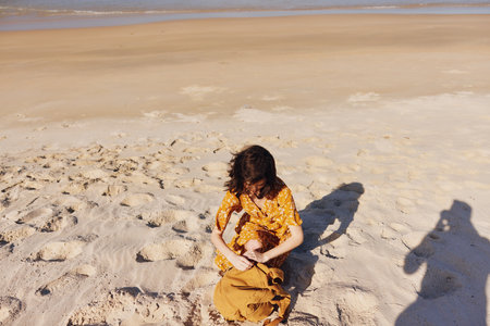 Woman in yellow dress sitting on beach with shadow stretching across sand on sunny dayの写真素材