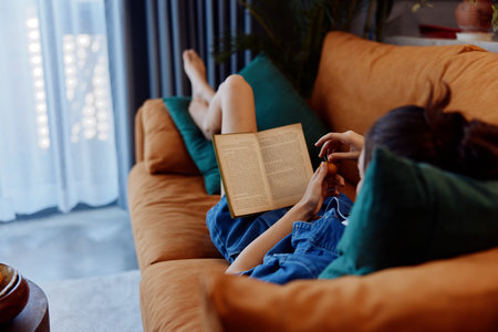 Woman relaxing on a couch by the window, engrossed in a book in her cozy living room at homeの写真素材
