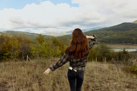 A woman enjoying the serene beauty of nature in a plaid shirt standing in a picturesque field with majestic mountains in the backgroundの写真素材
