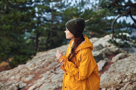 A woman in a yellow raincoat standing on a rocky hill with trees in the background, enjoying nature and adventureの写真素材