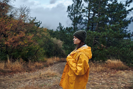 A young woman in a stylish yellow raincoat standing on a picturesque dirt road surrounded by lush...