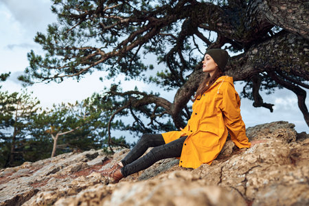 Woman in a yellow raincoat sitting on a rock in front of a tree during a peaceful nature retreatの写真素材