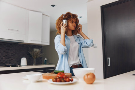 Young woman enjoying music with headphones in kitchen at home, African American female listening to music at homeの写真素材