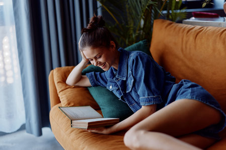 Young woman relaxing on couch with legs crossed reading an open book in cozy home environmentの写真素材