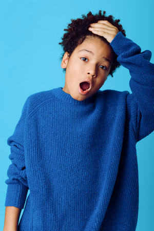 Surprised african american boy in blue sweater with hands on head against matching blue background expresses shock and amazementの写真素材