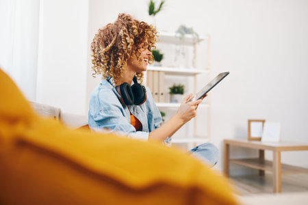 Home-reading Woman: A Happy, Smiling Gamer Girl Sitting on a Cozy Couch, Holding a Tablet PC, Enjoying Online Games and Chatting with a Friend via Headphones, Surrounded by a Beautifully Decorated Living Room.の写真素材