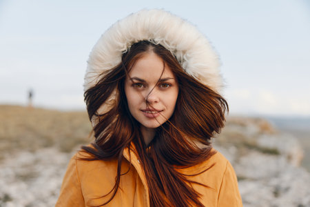 Woman in orange jacket and fur hat enjoying the beauty of nature on rocky hillside with blowing hairの写真素材