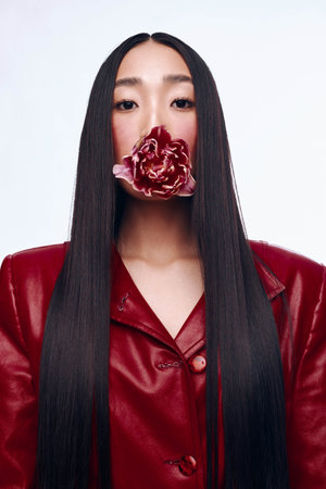 Elegant blackhaired woman in red jacket with flower in mouth posing for cameraの写真素材
