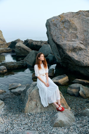 Woman in white dress relaxing on ocean rock with red shoesの写真素材