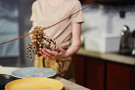 Woman holding lobster on plate in front of kitchen sink with bowl of food on counter, preparing seafood mealの写真素材