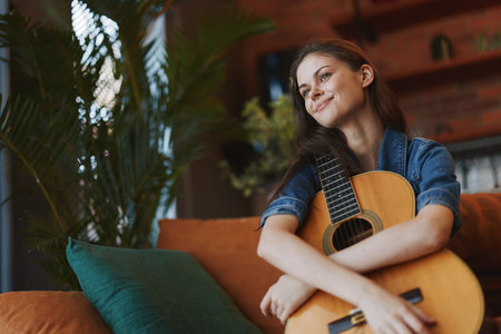 Young woman sitting on couch with acoustic guitar in front of window, enjoying peaceful moment of...