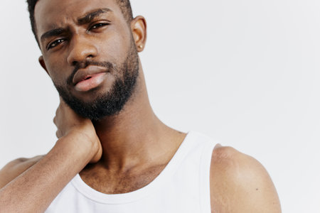 African american man in white tank top holding his neck in pain, experiencing discomfort or muscle...