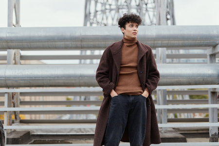 Fashion man in brown sweater, coat and black pants standing on a bridge with cityscape in the...
