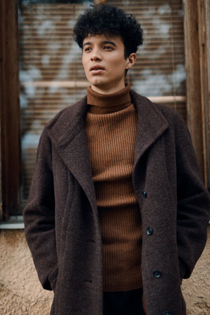 Fashionable young man in brown coat and turtleneck sweater standing in front of windowの写真素材