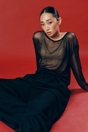Fashion asian woman in sheer top and pants sitting on red background with eyes closed and hands on...