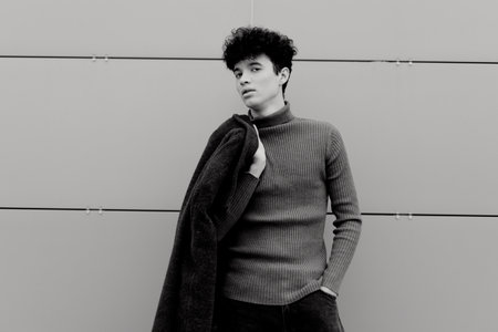 Fashionable black and white photo of a young man in a sweater and jeans leaning against a wall with his hands in his pocketsの写真素材