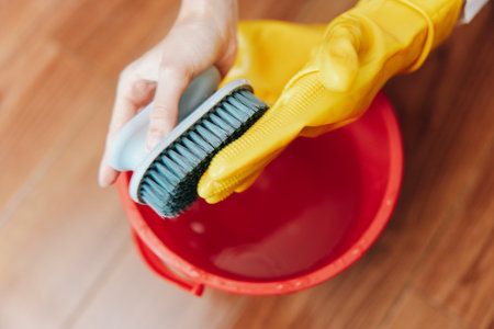Person in yellow rubber gloves cleaning red bucket with brush and water on wooden floorの写真素材