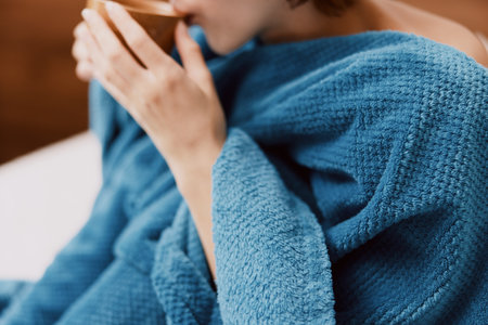 Woman wrapped in blue blanket sitting on bed in bedroom with cup of coffee in handの写真素材