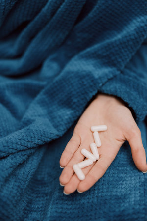 Person holds pills in hand wrapped in blue blanket with pillows next to them