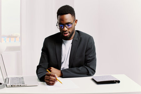Focused african american man in glasses working on laptop and taking notes at deskの写真素材
