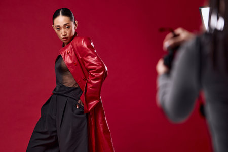 Fashionable asian woman in red leather coat and pants posing for a photographer in front of a red backgroundの写真素材