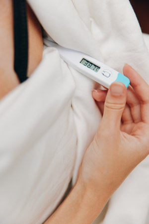Sick woman in bed holding electronic thermometer with blanket over head and pillow in front