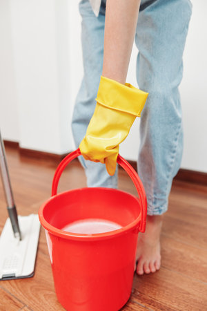 Woman with yellow rubber gloves holding a bucket of water on a hardwood floor with a mop in front of themの写真素材