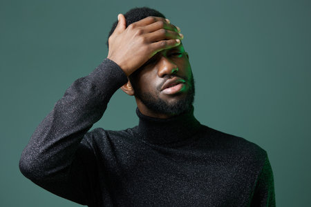Stressed bearded man in black sweater holding his head in frustration against a vibrant green...